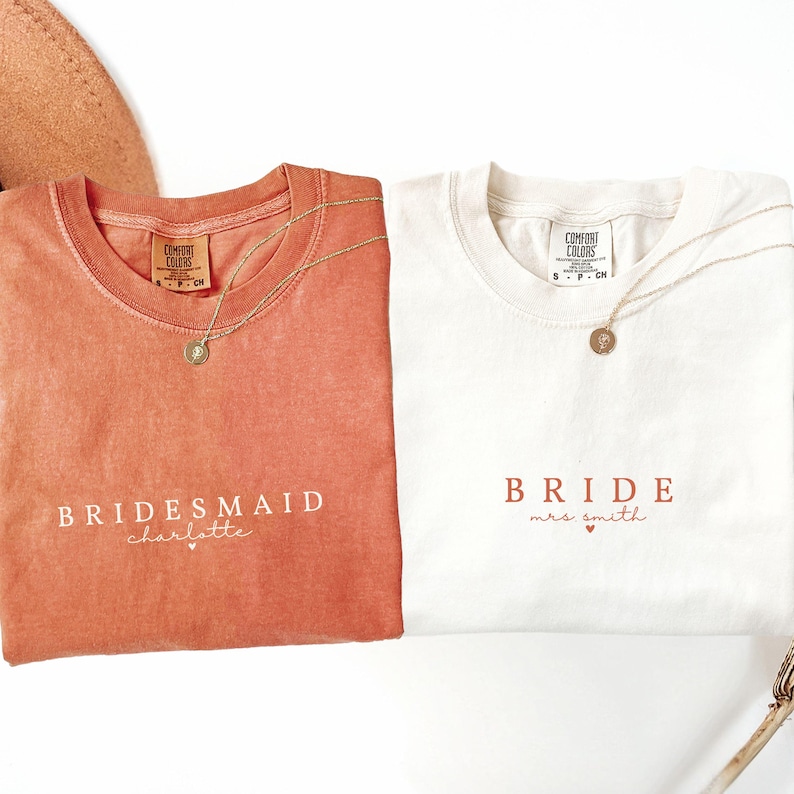 Puede incluir: Dos camisetas, una coral y otra blanquecina, con collares dorados. La camiseta coral dice "Bridesmaid Charlotte" en escritura blanca. La camiseta blanquecina dice "Bride Mrs. Smith" en escritura coral. Ambas camisetas tienen una etiqueta Comfort Colors.