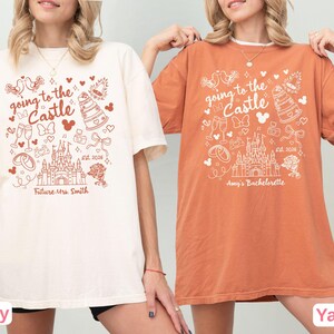 May include: Two t-shirts, one ivory and one yam-colored, feature a whimsical design with the text "going to the Castle." The design includes a castle, wedding rings, champagne glasses, and the text "Future Mrs. Smith" and "Amy's Bachelorette."