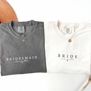 Puede incluir: Dos camisetas, una gris y otra blanca, con texto. La camiseta gris dice "Bridesmaid charlotte" y la camiseta blanca dice "Bride mrs. smith". Ambas camisetas tienen un collar de oro.