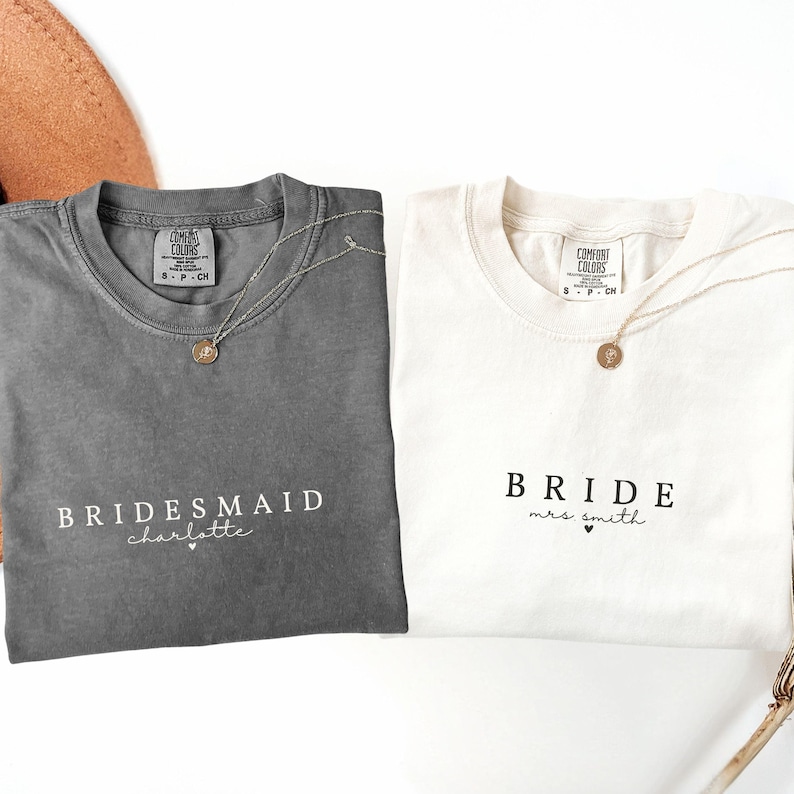 Puede incluir: Dos camisetas, una gris y otra blanca, con texto. La camiseta gris dice "Bridesmaid charlotte" y la camiseta blanca dice "Bride mrs. smith". Ambas camisetas tienen un collar de oro.