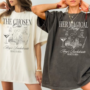 May include: Two oversized t-shirts, one cream and one charcoal, featuring a black graphic design. The design includes text, an owl, glasses, a wizard hat, a castle, a cocktail, and a broom. The text reads "THE CHOSEN One" and "HER MAGICAL Army".
