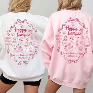 K&ouml;nnte beinhalten: Zwei Sweatshirts, eines wei&szlig; und eines rosa, jeweils mit einer rosa Grafik. Die Grafik lautet "Happy Camper" mit Illustrationen zum Thema Camping und "Carol's Bachelorette Scottsdale, AZ."