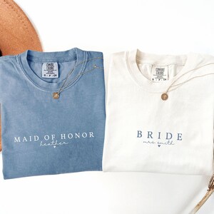 Puede incluir: Dos camisetas Comfort Colors, una azul y otra blanca, con collares dorados. La camiseta azul dice "MAID OF HONOR" y "heather". La camiseta blanca dice "BRIDE mrs. smith". Ambas camisetas tienen una etiqueta peque&ntilde;a.