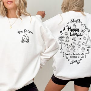 K&ouml;nnte beinhalten: Wei&szlig;es Sweatshirt mit schwarzen Grafiken. Auf der Vorderseite steht "The Bride" &uuml;ber einer Lagerfeuer-Illustration. Auf der R&uuml;ckseite steht "Happy Camper" mit Camping-Symbolen und "Carol's Bachelorette Scottsdale, AZ".