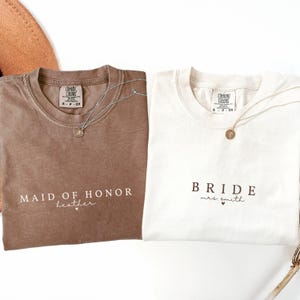 Puede incluir: Dos camisetas, una marr&oacute;n y otra blanca, con texto. La camiseta marr&oacute;n dice "MAID OF HONOR" y "heather". La camiseta blanca dice "BRIDE mrs. smith". Cada camiseta tiene un collar de plata.