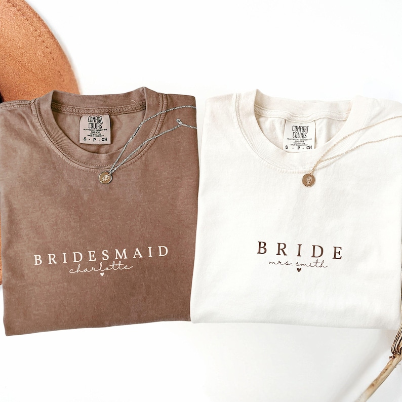 Puede incluir: Dos camisetas, una marr&oacute;n y otra blanca, con texto. La camiseta marr&oacute;n dice "Bridesmaid Charlotte" y la camiseta blanca dice "Bride Mrs. Smith". Ambas camisetas tienen un collar de plata con un colgante.