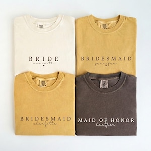 Puede incluir: Cuatro camisetas en crema, mostaza y carb&oacute;n. Cada camiseta tiene texto impreso. La camiseta crema dice "BRIDE mrs smith". Las camisetas mostaza dicen "BRIDESMAID jennifer" y "BRIDESMAID charlotte". La camiseta carb&oacute;n dice "MAID OF HONOR heather".