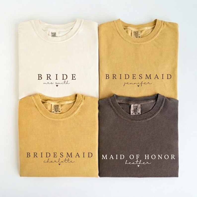 Puede incluir: Cuatro camisetas en crema, mostaza y carb&oacute;n. Cada camiseta tiene texto impreso. La camiseta crema dice "BRIDE mrs smith". Las camisetas mostaza dicen "BRIDESMAID jennifer" y "BRIDESMAID charlotte". La camiseta carb&oacute;n dice "MAID OF HONOR heather".