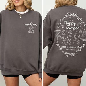 K&ouml;nnte beinhalten: Anthrazitfarbenes Sweatshirt mit wei&szlig;em Text und Grafiken. Auf der Vorderseite steht "The Bride" &uuml;ber einer Lagerfeuergrafik. Die R&uuml;ckseite zeigt ein "Happy Camper"-Design mit Camping-Symbolen und "Carol's Bachelorette Scottsdale, AZ".