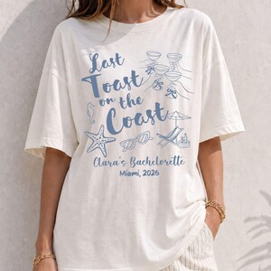 May include: An off-white short-sleeve t-shirt featuring blue graphic illustrations of hands holding wine glasses, a starfish, sunglasses, and a beach chair. The text reads Last Toast on the Coast, Clara's Bachelorette, and Miami, 2026.