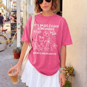 May include: A pink t-shirt with the text "IT'S BRIDE O'CLOCK SOMEWHERE" and "SARAH'S BACHELORETTE" in white. The shirt features white illustrations of a cocktail, rings, palm trees, and beach elements. The date "June 2026" is also printed on the shirt.