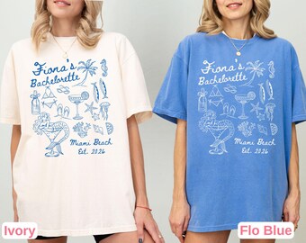 May include: Ivory and blue or Flo Blue t-shirts with a graphic design for a bachelorette party. The design includes the text "Fiona's Bachelorette", "Miami Beach Est. 2026", and various beach-themed illustrations like a mermaid, palm tree, and cocktails.