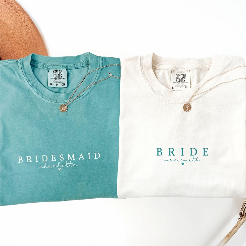 Puede incluir: Dos camisetas, una turquesa y otra blanca, con collares dorados. La camiseta turquesa dice "Bridesmaid Charlotte" en escritura blanca. La camiseta blanca dice "Bride Mrs. Smith" en escritura turquesa. Ambas camisetas tienen el logo "Comfort Colors".