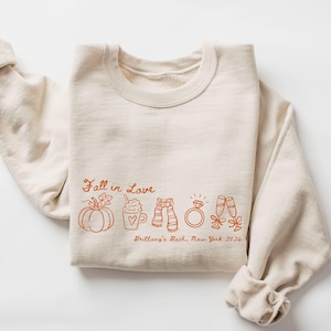 May include: A cream-colored crewneck sweatshirt featuring orange line art graphics of a pumpkin, a coffee mug, a scarf, a diamond ring, and two champagne flutes. The text reads Fall in Love and Brittany's Bach, New York 2026.
