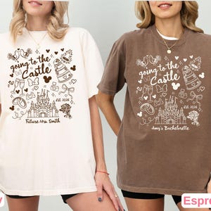 May include: Ivory and espresso-colored t-shirts with a whimsical design. The shirts feature the text "going to the Castle" with a castle illustration, Mickey Mouse ears, and other wedding-themed graphics. The ivory shirt says "Future Mrs. Smith" and the espresso shirt says "Amy's Bachelorette".