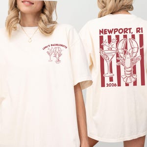 May include: An off-white short-sleeve t-shirt featuring a small red graphic on the front left chest with a lobster, martini glass, and the text LUNA'S BACHELORETTE. The back displays a large red graphic with vertical stripes, a lobster, a martini glass, and the text NEWPORT, RI 2026.