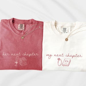 May include: A dark pink t-shirt with white text reading her next chapter and a champagne glass and book graphic, and a white t-shirt with dark pink text reading my next chapter and a ring and book graphic, each with a small gold necklace.