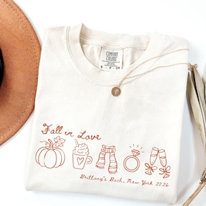 May include: A cream-colored t-shirt featuring the text Fall in Love and Brittany's Bach, New York 2026 in rust-colored ink. The graphic includes line art of a pumpkin, a mug with a heart, a scarf, a diamond ring, and two champagne flutes.