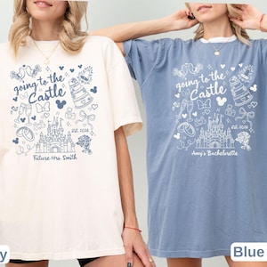 May include: Two t-shirts, one ivory and one blue, with a whimsical design. The shirts feature the text "Going to the Castle" with a castle illustration, doves, and other wedding-themed graphics. One shirt says "Future Mrs. Smith" and the other says "Amy's Bachelorette".