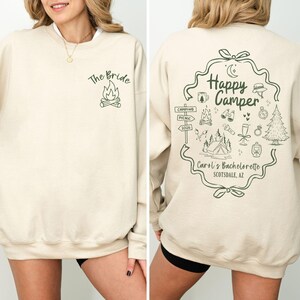 K&ouml;nnte beinhalten: Cremefarbenes Sweatshirt mit dem Text "The Bride" und einer Lagerfeuer-Grafik auf der Vorderseite. Die R&uuml;ckseite zeigt ein "Happy Camper"-Design mit Camping-Illustrationen und dem Text "Carol's Bachelorette Scottsdale, AZ."