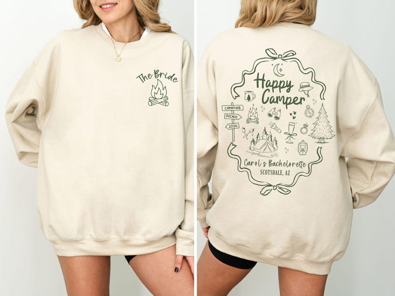 K&ouml;nnte beinhalten: Cremefarbenes Sweatshirt mit dem Text "The Bride" und einer Lagerfeuer-Grafik auf der Vorderseite. Die R&uuml;ckseite zeigt ein "Happy Camper"-Design mit Camping-Illustrationen und dem Text "Carol's Bachelorette Scottsdale, AZ."