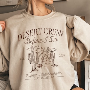 May include: Beige crewneck sweatshirt with brown text and a desert-themed graphic. The text reads "DESERT CREW Before I Do" with "Sophia's Bachelorette" and "Scottsdale, AZ" below a desert landscape illustration. The year "2026" is also printed.