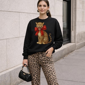 Può includere: Felpa nera con un leopardo con un fiocco rosso. Il leopardo ha un motivo maculato ed è seduto. La felpa è abbinata a jeans con stampa leopardata. Una borsa nera è tenuta in mano.