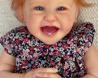 May include: A realistic baby doll with ginger hair tied with a yellow pineapple clip, blue eyes, and a wide smile showing one tooth. The doll wears a navy blue dress with a pink and white floral pattern, gold earrings, and a gold charm bracelet.