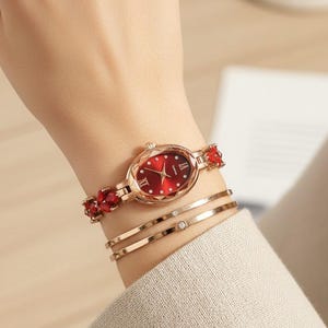 Può includere: Un orologio color oro rosa con quadrante rosso e numeri romani. Il cinturino dell'orologio presenta dettagli di pietre preziose rosse. Sono visibili anche due braccialetti in oro rosa con piccoli accenti di diamanti.