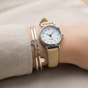 Puede incluir: Un reloj de pulsera con esfera blanca y agujas negras, con una correa de cuero beige. El reloj está en una muñeca con pulseras plateadas. La esfera del reloj tiene la palabra "Quartz" impresa.
