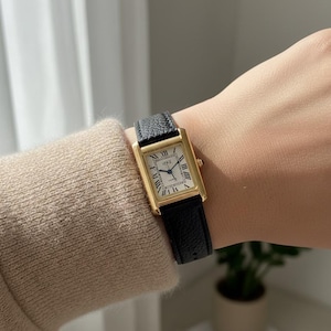 Pode incluir: Um relógio de pulso retangular dourado com uma pulseira de couro preta. O mostrador do relógio é branco com algarismos romanos pretos e ponteiros dourados. O relógio está em um pulso.