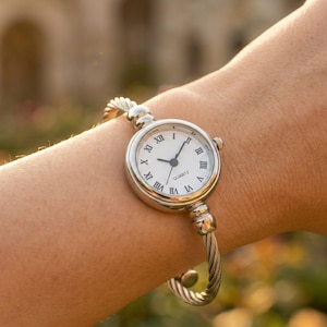 Może przedstawiać: Srebrny zegarek kwarcowy z białą tarczą i czarnymi rzymskimi cyframi jest noszony na nadgarstku. Zegarek ma okrągłą tarczę i skręconą metalową bransoletę. Zegarek jest w klasycznym stylu.