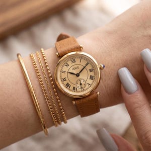 May include: A gold-toned wristwatch with a tan leather strap is displayed on a wrist, alongside gold bracelets. The watch face features Roman numerals and the brand name "IEKE". The watch is approximately 3.8 cm in diameter.