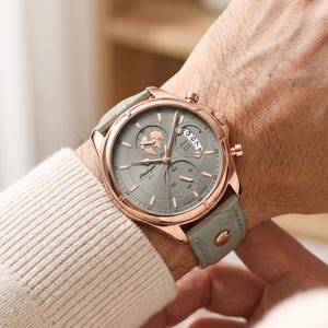 Puede incluir: Un reloj de pulsera de oro rosa con esfera gris y correa de cuero gris. La esfera del reloj presenta múltiples esferas y la marca "Pindagar 1853". El reloj está en una muñeca.