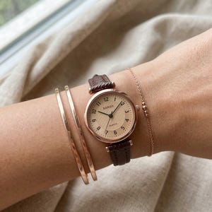 May include: A rose gold watch with a brown leather strap and a cream-coloured face. The watch face displays the word "BARSH". Two rose gold bangles and a delicate rose gold bracelet are also visible.