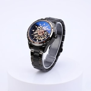 May include: Black skeleton watch with a stainless steel strap. The watch face has a blue tint and a visible mechanical movement with gold-coloured gears. The watch is displayed on a white surface.