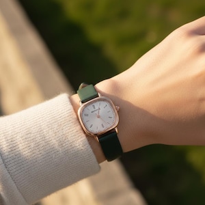 Könnte beinhalten: Eine roségoldene Armbanduhr mit quadratischem Zifferblatt und weißem Zifferblatt. Die Uhr hat ein dunkelgrünes Lederarmband. Die Uhr befindet sich an einem Handgelenk, wobei ein hellfarbiger Pulloverärmel sichtbar ist.