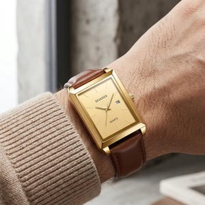 Könnte beinhalten: Eine goldfarbene rechteckige Uhr mit einem braunen Lederarmband. Das Zifferblatt der Uhr ist goldfarben mit schwarzen Zeigern und dem Wort "DIJANES" oben aufgedruckt. Die Uhr hat auch eine Datumsanzeige. Es ist eine Quarzuhr.