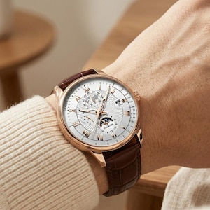 May include: A rose gold wristwatch with a white face and brown leather band. The watch face features multiple subdials and a moon phase indicator. The band is a dark brown color and has a textured appearance.