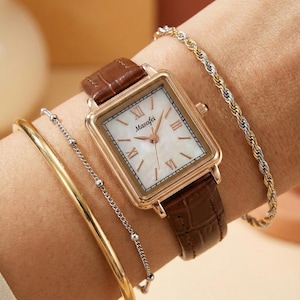 Puede incluir: Un reloj rectangular dorado con una correa de cuero marrón se muestra en una muñeca, acompañado de pulseras doradas y plateadas. La esfera del reloj es blanca con números romanos y la marca "Masafei".