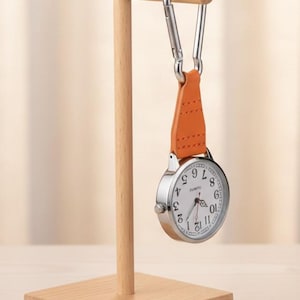 Op de afbeelding: Een zilverkleurig zakhorloge met een witte wijzerplaat en zwarte cijfers hangt aan een oranje leren band en een zilveren karabijnhaak. Het horloge wordt getoond op een kleine houten standaard.