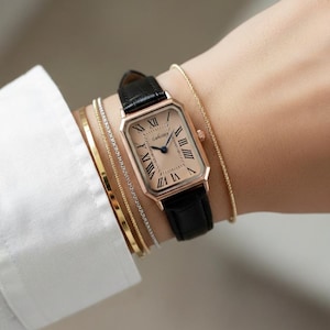 Puede incluir: Un reloj rectangular de oro rosa con una correa de cuero negro. La esfera del reloj es de color melocotón claro con números romanos negros. Se usan varias pulseras doradas y plateadas.