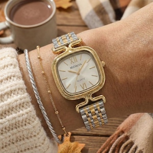 May include: A gold and silver wristwatch with a square face is displayed. The watch face is white with gold Roman numerals and the brand name "MIDAIYA". The watch is on a wrist with a silver and gold bracelet.