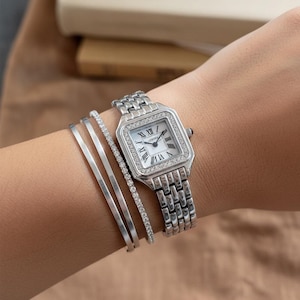 May include: A silver wristwatch with a square face and a diamond-set bezel. The watch features a white dial with Roman numerals and a silver link bracelet. Two silver bangles and a diamond bracelet are also visible.