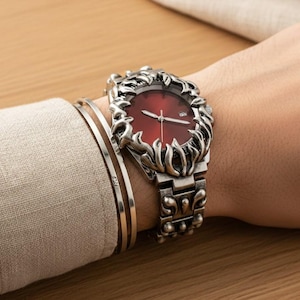 Peut inclure: Une montre argentée avec un cadran rouge et des détails décoratifs en forme de flammes autour de la lunette. La montre est sur un bracelet argenté assorti. Deux bracelets argentés sont également visibles.