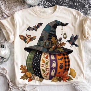 May include: A cream-colored t-shirt featuring a graphic of a pumpkin wearing a patchwork witch hat. The pumpkin is decorated with patterns of ghosts, owls, bats, leaves, and stars. A bat, an owl, and a crow surround the hat, which is adorned with a key and a lantern.