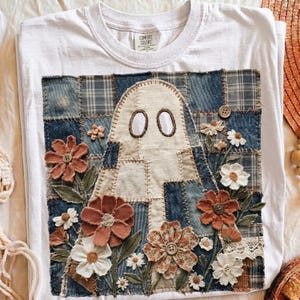May include: A white t-shirt featuring a large rectangular patch made of blue denim and plaid fabric squares. The patch displays a central ghost figure surrounded by fabric flowers in shades of rust, white, and brown, accented with small buttons.