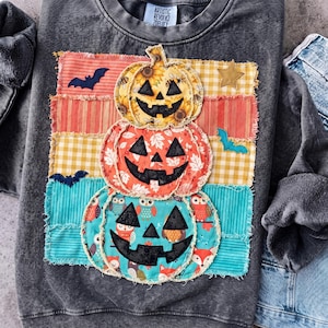 May include: A charcoal gray acid-wash sweatshirt featuring a stacked pumpkin applique design. The pumpkins are made of patterned fabric with black stitched faces. The background consists of horizontal fabric strips with bats and a star.