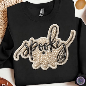 May include: A black crewneck sweatshirt featuring a central patch shaped like a ghost. The ghost is embellished with cream-coloured faux pearls and black beads, forming the word "spooky" in cursive script with a spiderweb detail on the side.