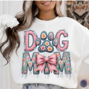 以下が含まれることがあります： ピンクとフローラルのデザインで「DOG MOM」の文字が入った白いスウェットシャツ。文字はデニムのような素材で縁取られ、花とピンクのリボンで飾られています。